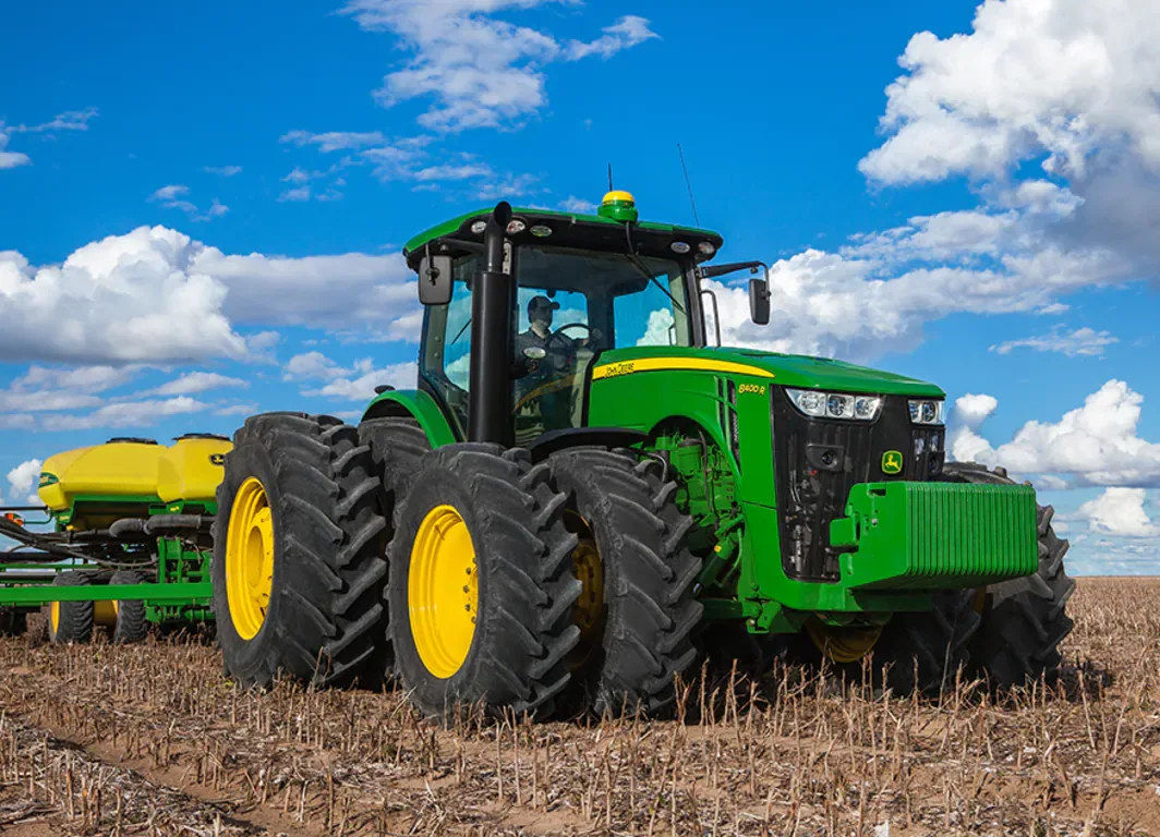 Trator John Deere 8400R verde e amarelo em um campo arado sob um céu azul, puxando um plantador amarelo.