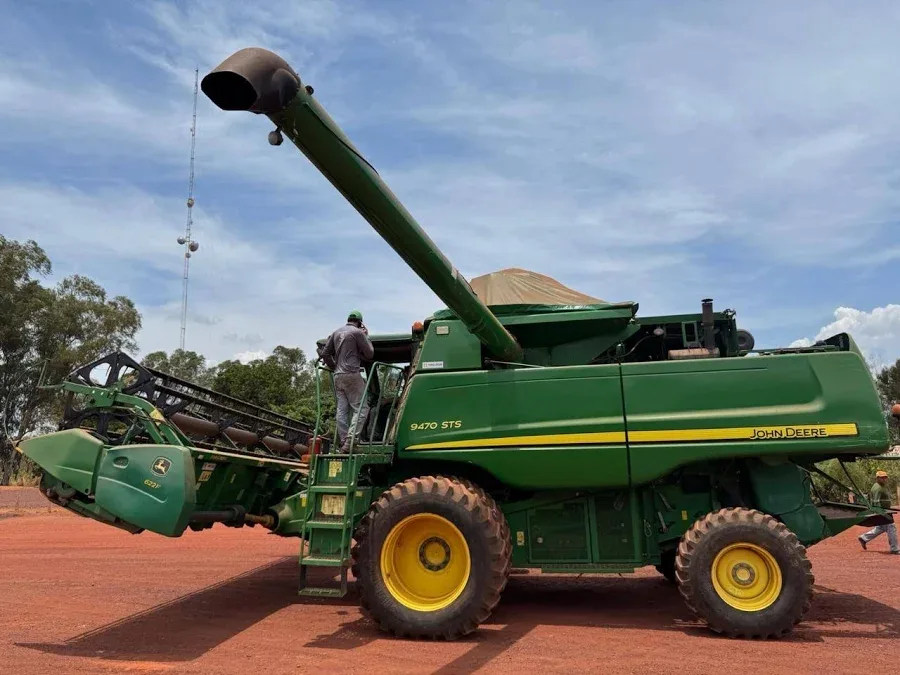 Imagem lateral completa de uma colheitadeira John Deere 9470 STS verde e amarela em um campo de terra batida.